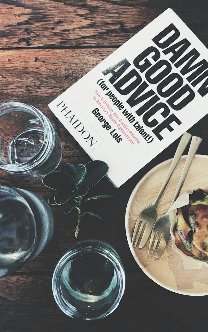 Ein Buch mit dem Titel "Damn Good Advice" liegt neben Gläsern und einem Gericht auf einem Holztisch.