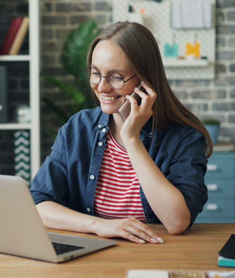 Frau mit Telefon in der Hand, sitzt am Schreibtisch und arbeitet am Laptop.