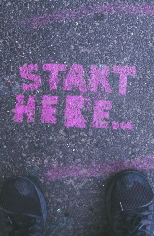 Pink "START HERE" Schriftzug auf dem Boden, sichtbar vor einem Paar Schuhe.